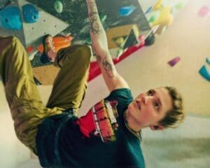 http://A%20young%20person%20with%20tattoos%20climbing%20an%20indoor%20rock%20wall%20with%20colorful%20holds,%20looking%20up%20intently.