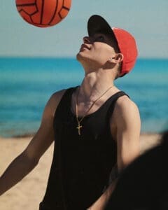 http://Young%20man%20in%20a%20black%20tank%20top%20and%20red%20cap%20looking%20up%20at%20an%20orange%20ball%20on%20a%20sunny%20beach%20with%20turquoise%20water.