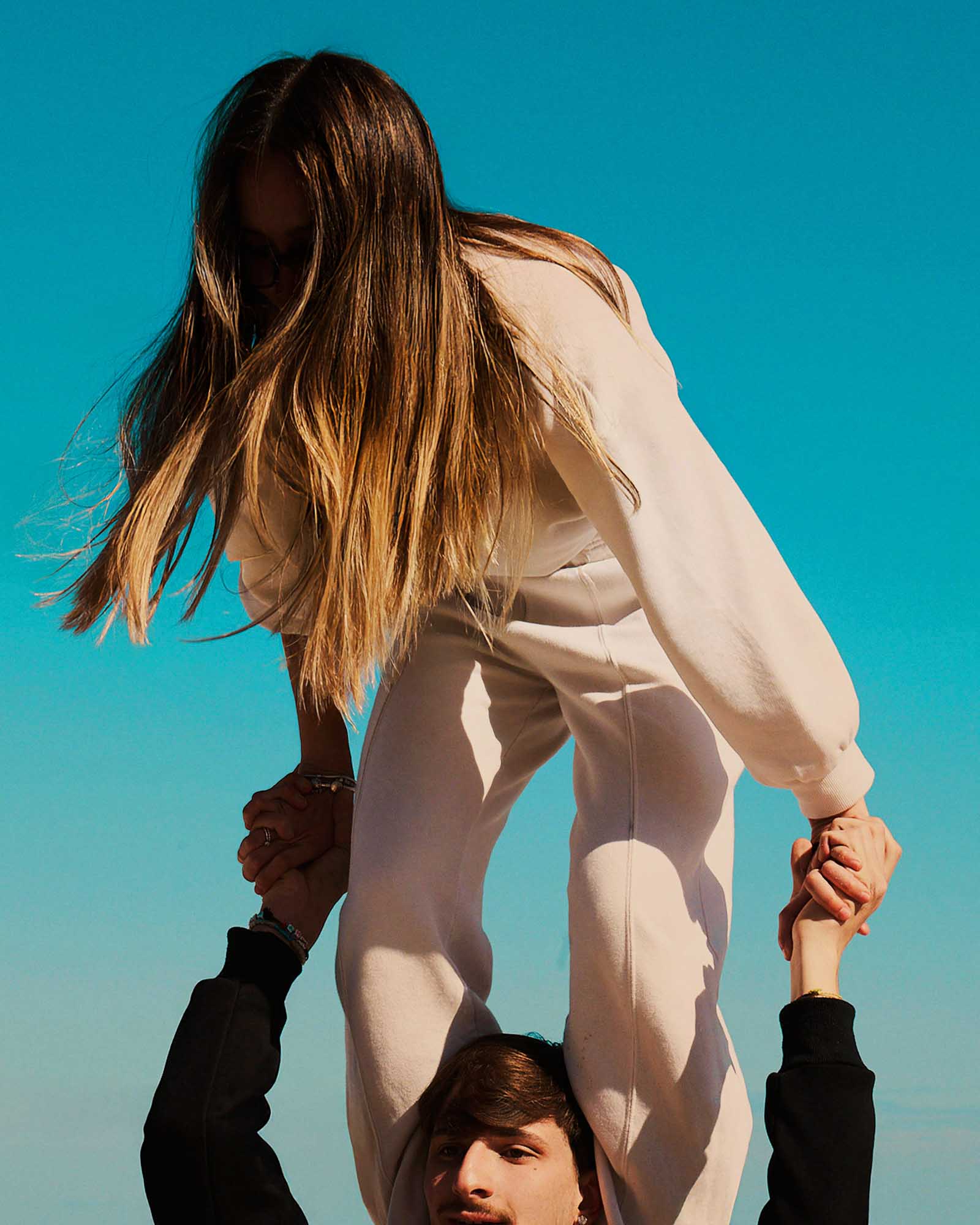 A low-angle shot features a young woman with long, light brown hair, wearing a light-colored sweatsuit, sitting on the shoulders of a young man. Her face is obscured by her hair. She holds his hands for balance, and his face is partially visible at the bottom of the frame. The background is a vibrant, clear turquoise sky, suggesting a sunny day.