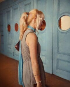 http://Candid%20profile%20photo%20of%20a%20young%20woman%20with%20pink%20hair%20in%20a%20high%20ponytail,%20wearing%20a%20gray%20dress%20and%20blue%20scarf,%20standing%20in%20front%20of%20light%20blue%20doors%20with%20circular%20windows.%20From%20the%20'In%20Between'%20project.