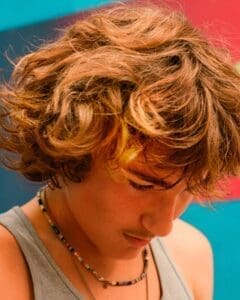 http://Close-up%20of%20a%20young%20person%20with%20wavy,%20sun-kissed%20brown%20hair%20looking%20down,%20wearing%20a%20grey%20tank%20top%20and%20beaded%20necklaces,%20against%20a%20colourful,%20blurred%20background.