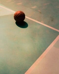 http://A%20single%20orange%20basketball%20resting%20on%20a%20pale%20teal%20outdoor%20court%20with%20white%20lines,%20casting%20a%20soft%20shadow.
