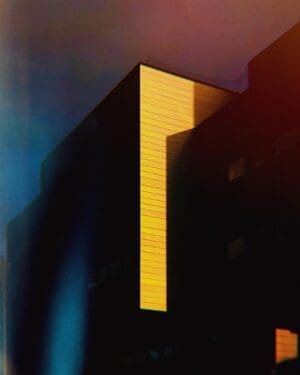 Abstract architectural detail of a building with a bright yellow vertical section against dark shadowed areas and a moody blue/red sky